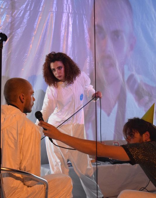 Performance artists wearing plastic jumpsuits and talking in a microphone. 
