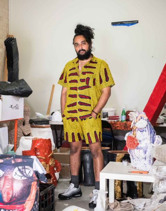 Ramesh Mario Nithiyendran in his studio. Artist in yellow outfit standing amongst artwork and materials.