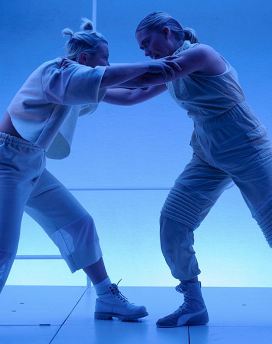Two people stand with their hands on each others shoulders, both dressed entirely in white. They stand on a blue lit stage.
