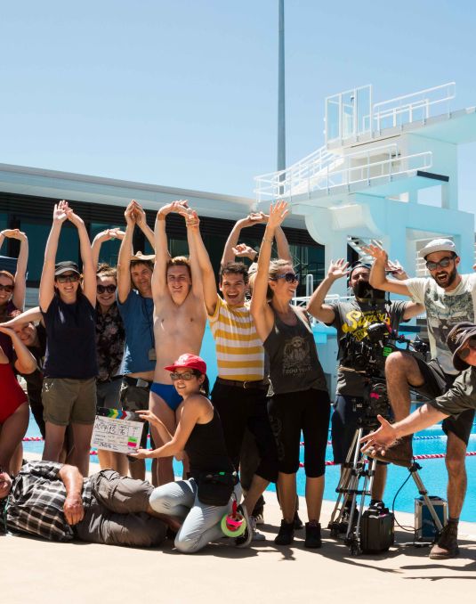 A large group of people stand at the side of an outdoor pool. Some hold filming equipment and some pose with their hands above their head. 