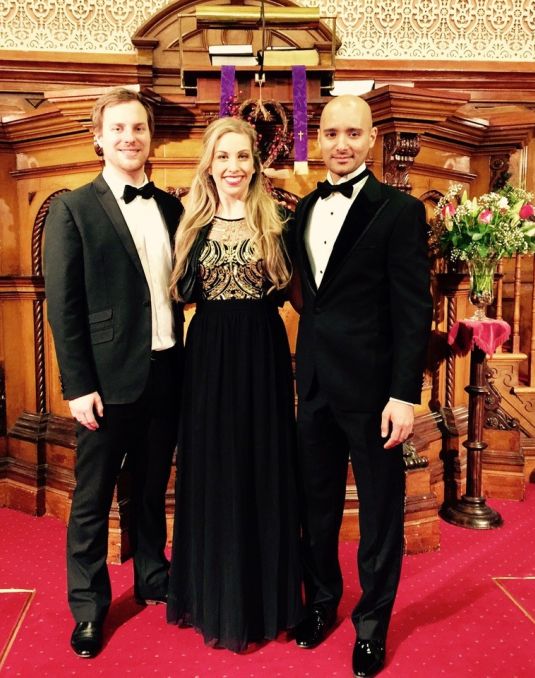 To men in tuxedos and woman in a floor length black gown pose on a red carpet.