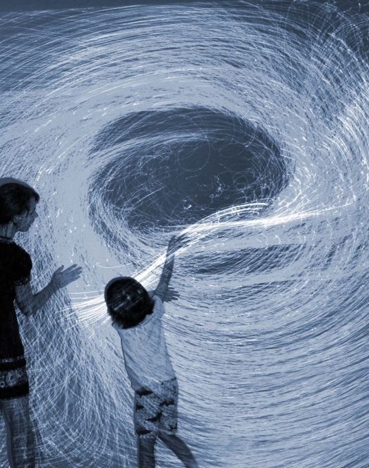 Two young children standing in front of a wall with a spiral of white light projected onto it.