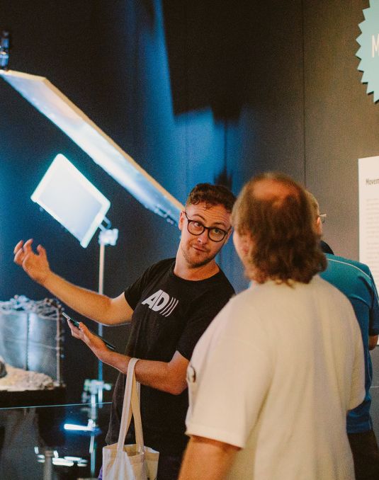 People looking at exhibition of stop start motion animation Wallace and Gromit. A man talks to another audience member. He wears a black t-shirt with the initials AD on the chest.