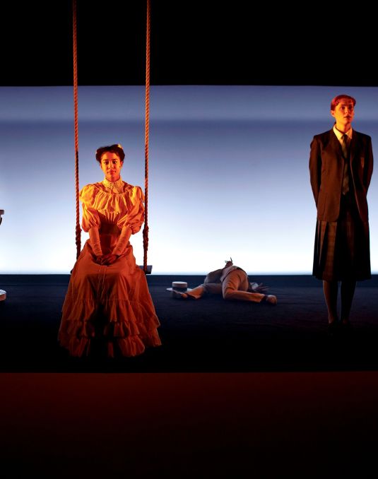 Moody still of theatre piece with a fountain, a woman on a swing and a man in a trenchcoat.
