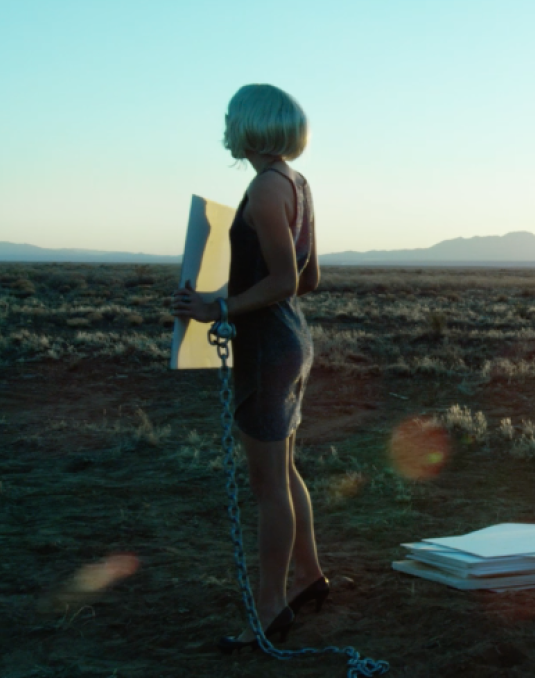 An actress stands on a grassy dessert landscape near a wire fence that stretches into the distance. The sun sets in the background.  