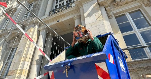 Artist holding onto artistic mask face covering emerging out of dumpster in front of building.