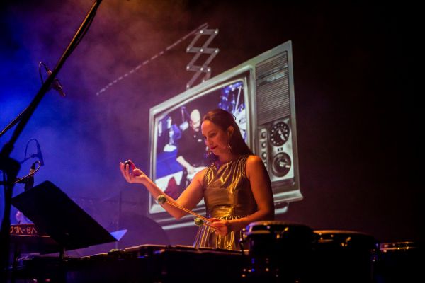 Woman performing on stage with xylophone and television projection in background.