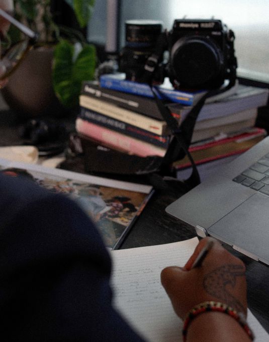 Artist's handwriting notes on desk with a camera and laptop perched nearby.