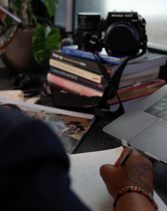 Artist's handwriting notes on desk with a camera and laptop perched nearby.