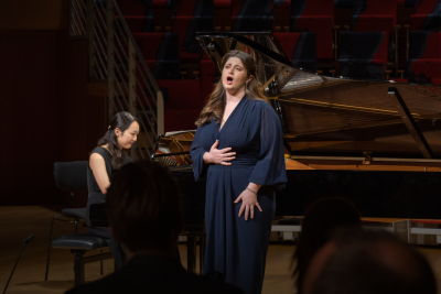 Woman in naxy dress singing to crowd, with woman playing piano in the background.