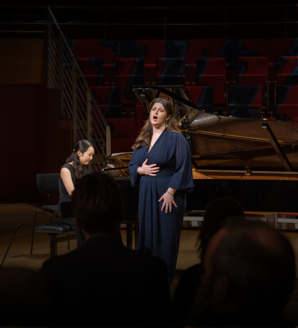 Woman in naxy dress singing to crowd, with woman playing piano in the background.