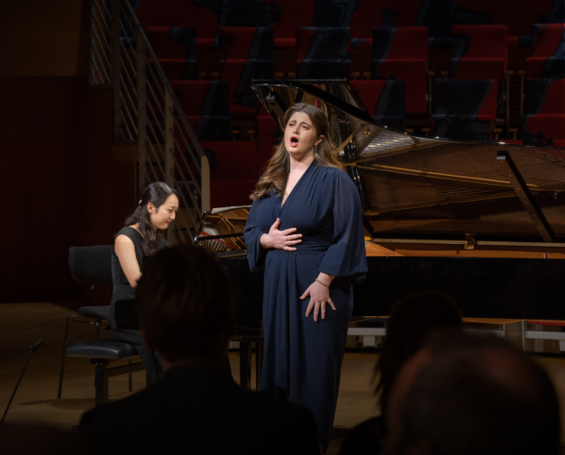 Woman in naxy dress singing to crowd, with woman playing piano in the background.