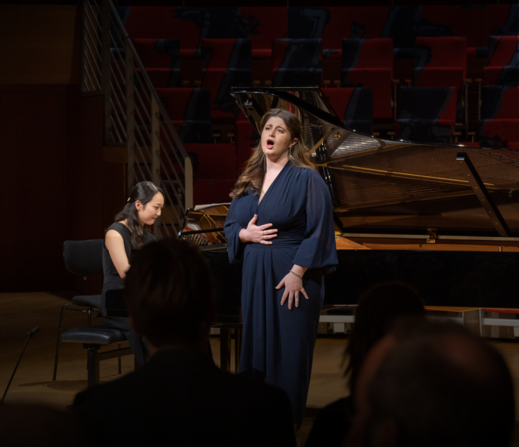Woman in naxy dress singing to crowd, with woman playing piano in the background.